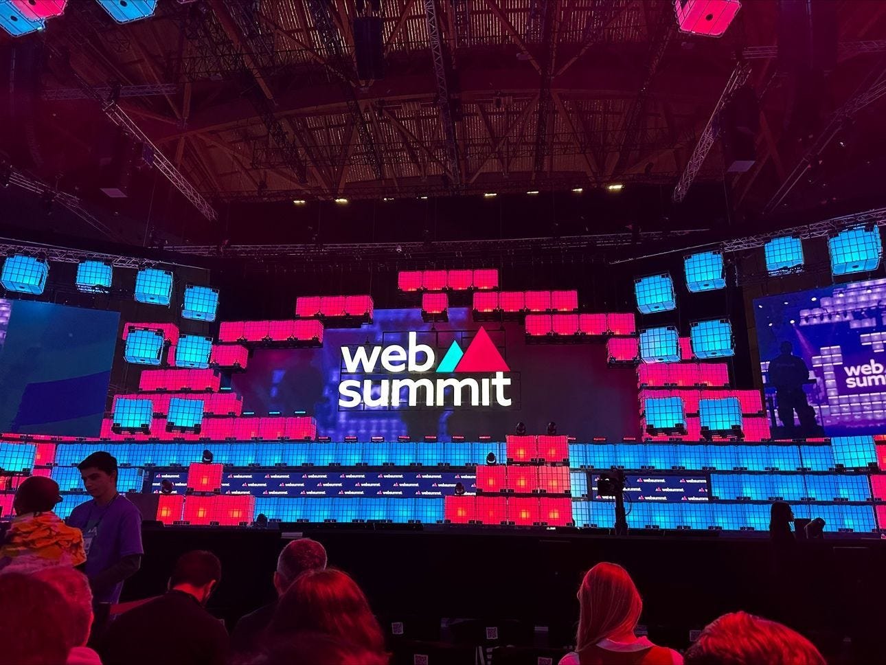 Palco principal do Web Summit iluminado a rosa e azul com participantes sentados à frente